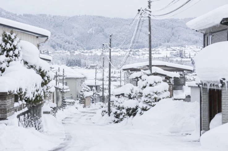 屋根や道路脇に雪が積もった住宅地と、奥に見える雪に覆われた山並み