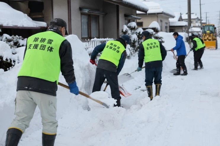 蛍光色のベストを着た人たちが、雪の積もった住宅地でスコップを使って除雪する様子