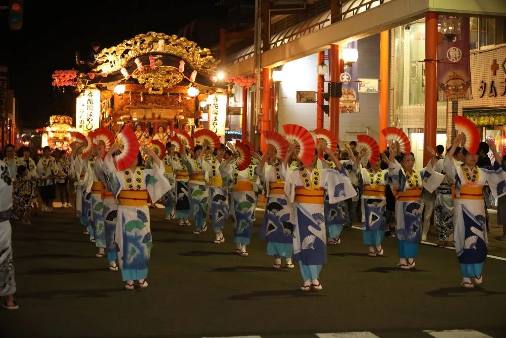 赤と白の扇を掲げて踊る、そろいの衣装を着た参加者たちと後ろを進む屋台