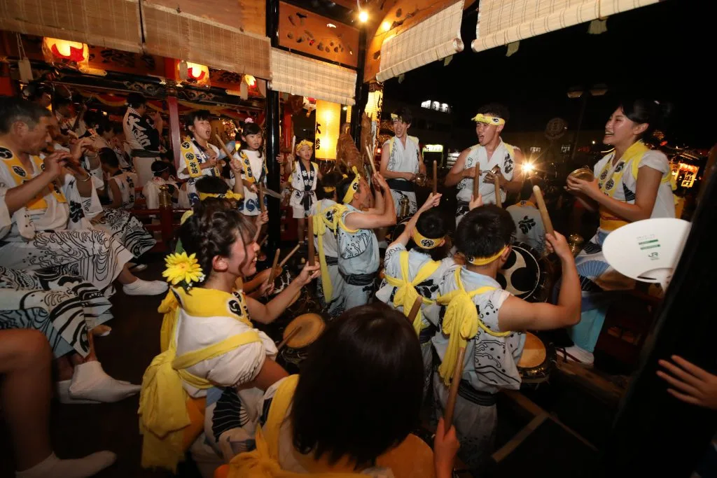 屋台の中で太鼓を囲み、ばちを持って演奏する花輪ばやしの参加者たち